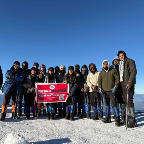 Group of happy trekkers posing with banner on snow-covered Kedarkantha peak during winter trek in Uttarakhand