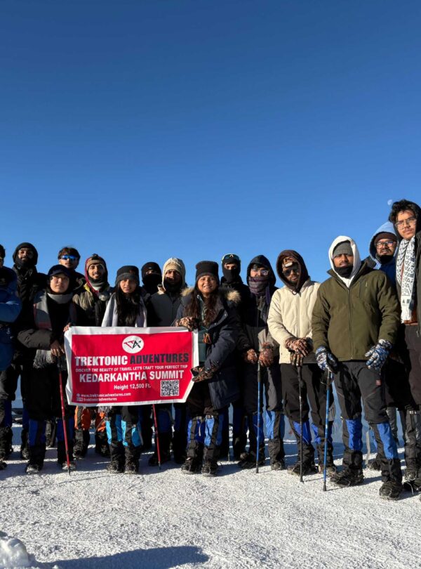 Group of happy trekkers posing with banner on snow-covered Kedarkantha peak during winter trek in Uttarakhand